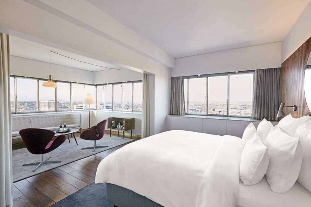 Quarto do Radisson Collection Royal Hotel com uma cama de casal, mesinhas e cadeiras e janelas panorâmicas com vista para a cidade. Foto para ilustrar post sobre hotéis em Copenhague.