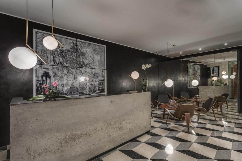 recepção do Navona Theatre Hotel com chão xadrez, bancada cinza, luzes redondas pendendo do teto e mesa com cadeiras em frente à um grande espelho estilizado