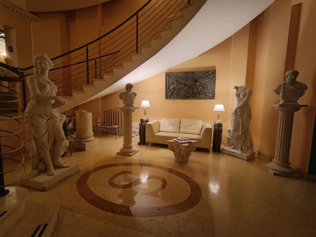 recepção do Seccy Hotel Boutique Art & Museum com a logo no chão, sofá branco com luminária dos lados, escadaria e diversas esculturas brancas em estilo clássico romano
