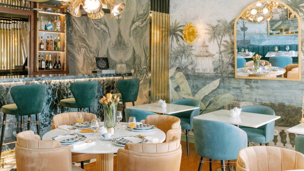 Restaurante luxuoso com bar ao fundo, mesas de mármore branco, cadeiras aveludadas verdes e beges, paredes com pinturas e espelho com moldura dourada.