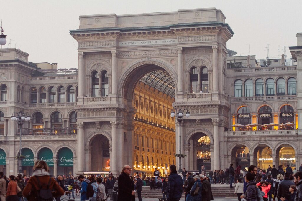 Galeria Vittorio Emanuele II uma construção histórica muito grande a alta com teto de vidro e sua entrada forma um arco, há muitas pessoas na frente do local