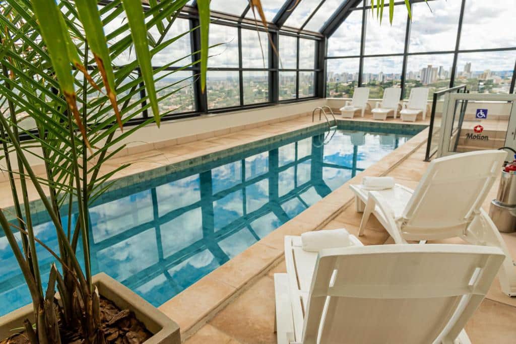 Área da piscina com cobertura em vidro, uma palmeira verde e algumas cadeiras brancas em volta durante o dia, ilustrando post Hotéis em Maringá.