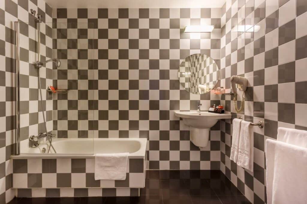 Banheiro com azulejos quadriculados no Room Mate Mario. Uma banheira com box encara uma pia com secador de cabelo, amenidades de banho e espelho. Há suportes com toalhas brancas nas paredes.