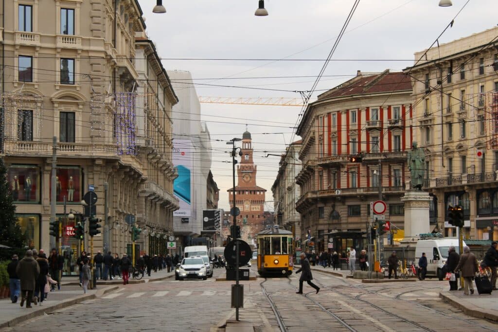 Rua movimentada de Milão com carros passando bondinhos amarelos, pessoas caminhando, os prédios ao redor são antigos e históricos