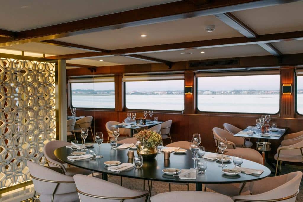 Mesas com pratos, copos, taças, guardanapos, flores, cadeiras e janelas com vista para o rio em restaurante de um barco hotel.