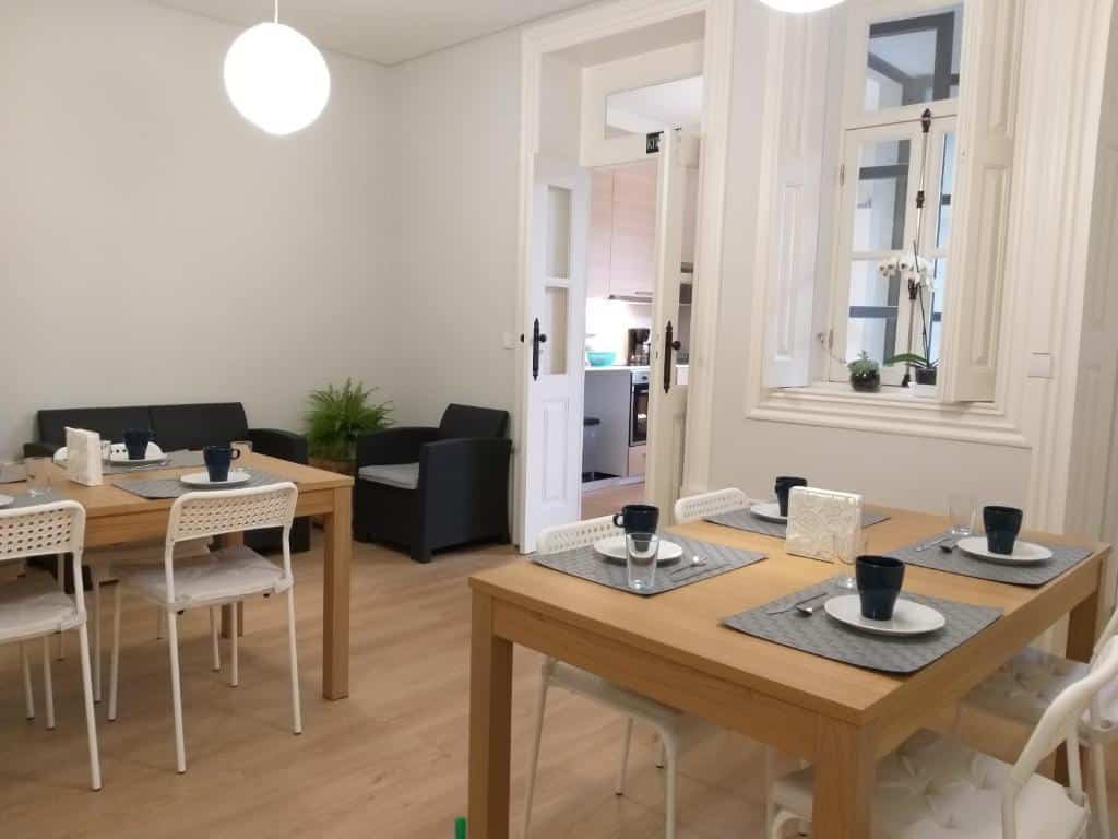 Sala de estar e de refeição do Queen’s Garden Hostel com duas mesas de madeira a frente com cadeiras e ao fundo sofá e uma poltrona.