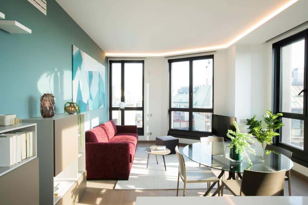 Sala de estar ddo Milan Eleven by Brera Apartments com três janelas amplas, um rack com televisão, uma pequena mesa de centro, um sofá vermelho e uma mesa redonda de vidro com quatro lugares