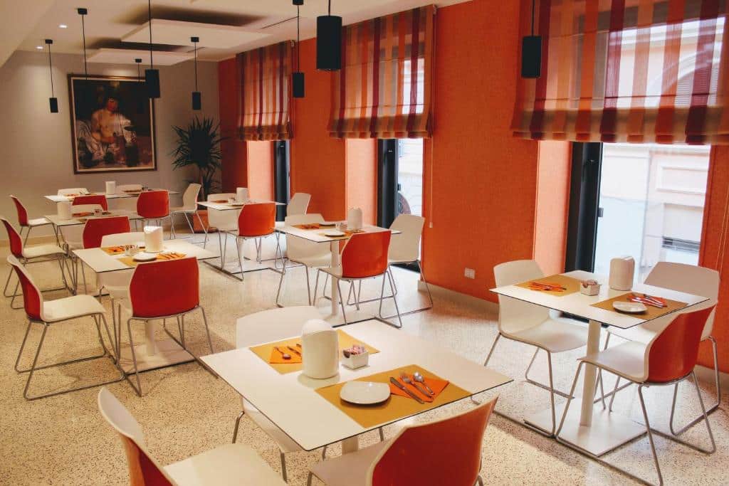 salão do Domus Helena com mesas quadradas brancas e cadeiras confortáveis laranjas combinando as paredes, há uma tela na parede e luminárias pretas que descem do teto