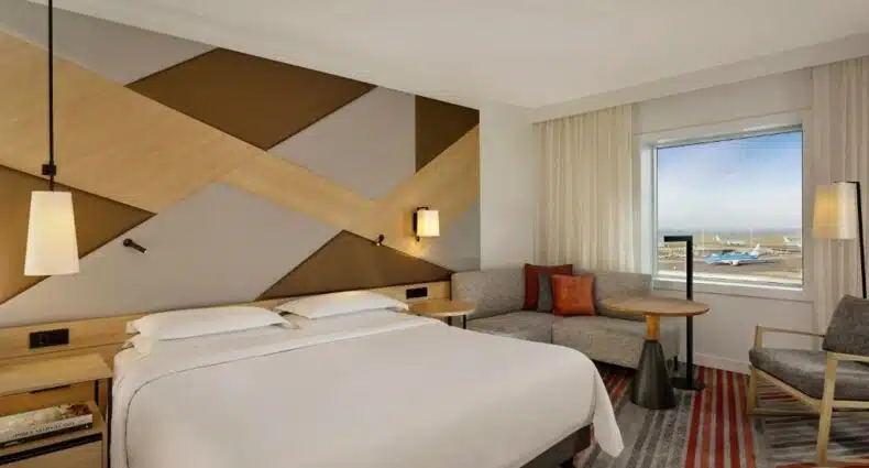 Quarto do Sheraton Amsterdam, com cama de casal, sofá de canto na parede esquerda, uma mesinha redonda e uma cadeira ao lado. Há, também, uma janela com vista para o aeroporto e avião. Imagem para ilustar o post de hotéis perto do aeroporto de Amsterdam