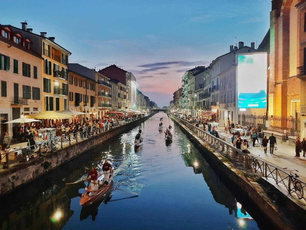 Um dos canais de Navigli, em Milão, com alguns barcos passando, e nas margens há muito comércio e pessoas andando