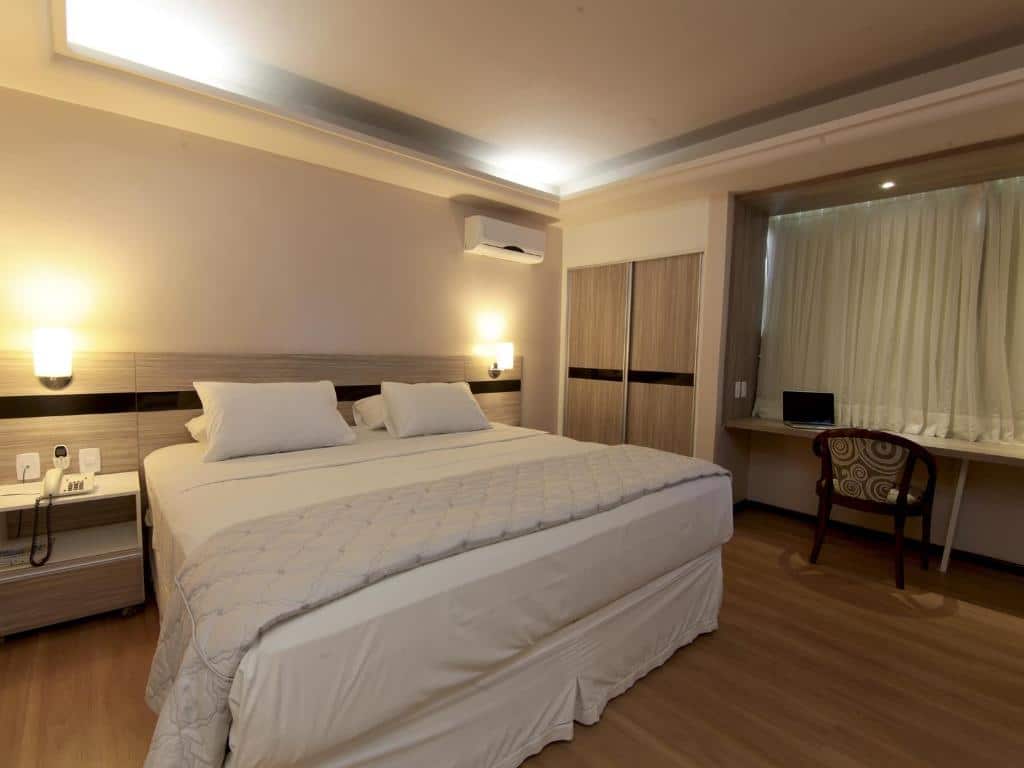 Quarto de hotel com cama de casal com colcha branca, móveis em madeira, pequena mesinha ao lado da cama, luminárias nos dois lados da cama e ar-condicionado. Imagem para ilustrar o post hotéis em Campina Grande.