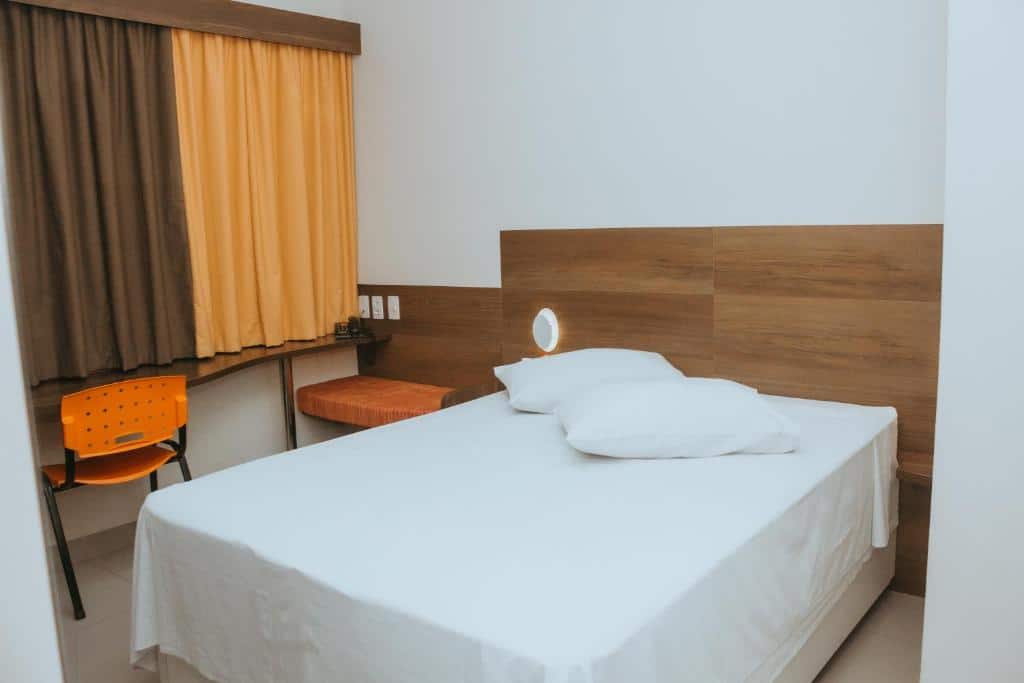 Quarto simples com cama de casal com lençol branco, cabeceira de madeira, mesa e cadeira ao lado da cama e cortina marrom e laranja. Imagem para ilustrar o post hotéis e pousadas em Itu.