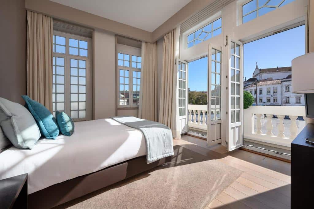 Quarto com cama de casal com colcha branca e travesseiros azuis, portas abertas mostrando uma varanda com vista. Imagem para ilustrar o post hotéis em Aveiro.