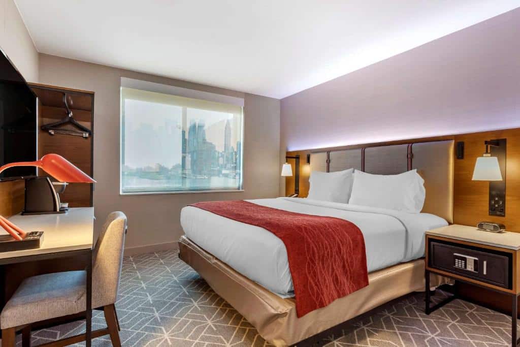 quarto do hotel Comfort Inn perto do aeroporto JFK em Nova York com uma cama de casal à direita, um cofre na escrivaninha, uma mesa para trabalho em frente a cama e uma janela ao fundo.