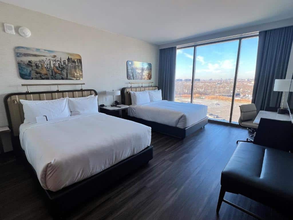 suíte dupla do Hyatt Regency JFK com duas camas de casal dispostas lado a lado no canto esquerdo da imagem, com uma parede de vidro ao fundo quarto mostrando toda a paisagem de Nova York.