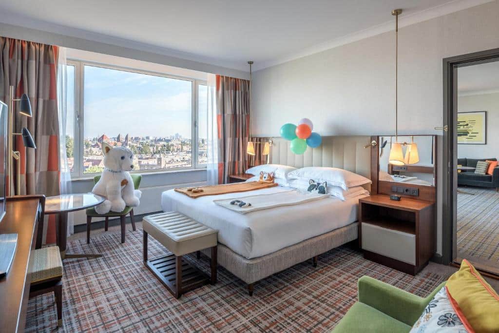 Suíte família do Hilton Amsterdam, de 36 m², com cama de casal com toalhas temática de ursos em cima, bexigas coloridas amarradas na cabeceira, e um urso em cima de uma cadeira. Há, ainda, uma janela grande com vista da cidade, espelho e uma cadeira estofada. Representa hotéis para família em Amsterdam