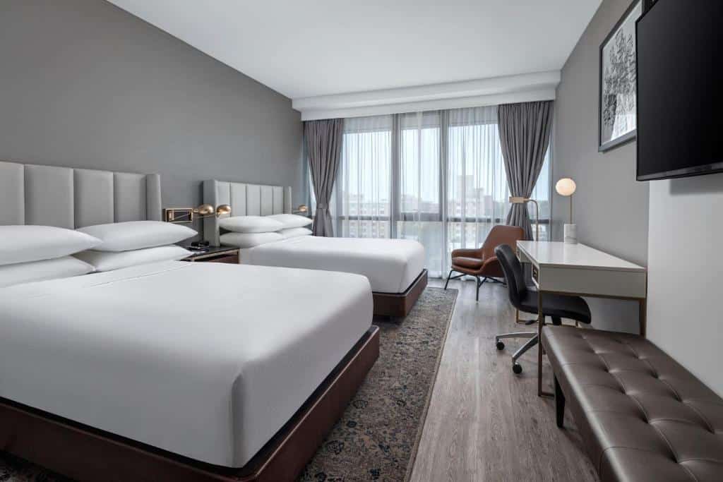 quarto do Four Points by Sheraton Flushing com duas camas de casal lado a lado com lençóis brancos e uma mesa branca em frente com uma cadeira preta de rodinhas
