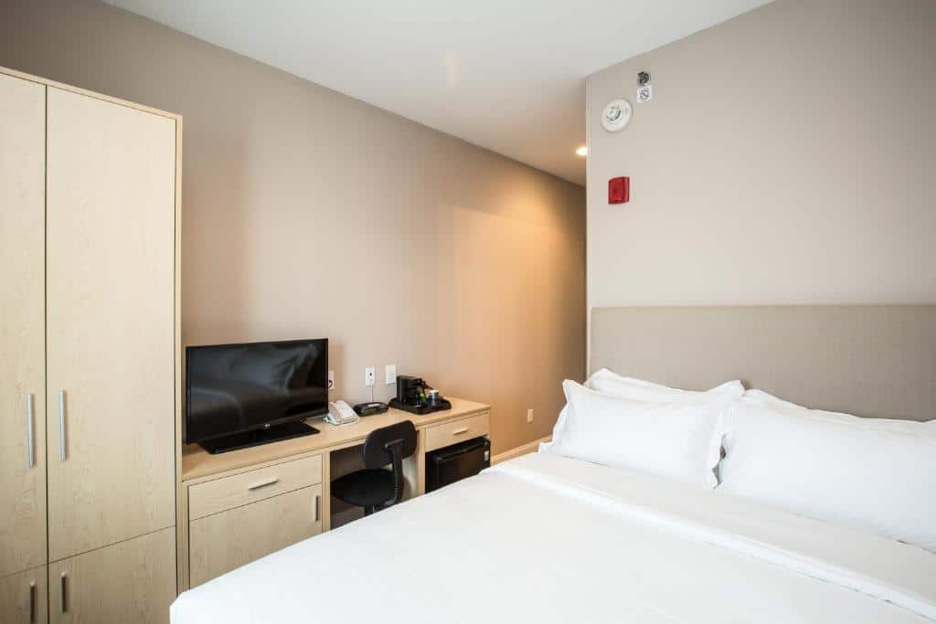 suíte do Grandview Hotel New York, um dos hotéis perto do aeroporto laguardia em nova york, com uma cama de casal à direita e um armário claro, de madeira, à esquerda, acompanhado de uma televisão pequena e uma mesa para trabalho