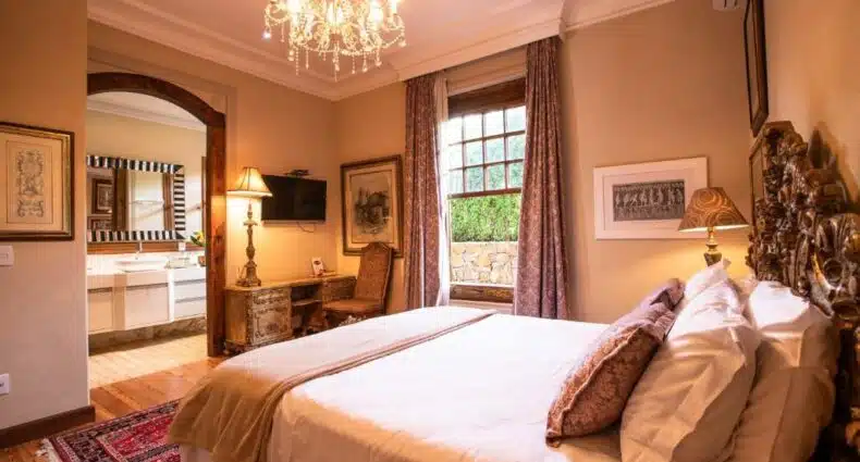 Imagem de uma das suítes da Pousada Villa Maria. A decoração é extremamente elegante, há um lustre no quarto, os móveis são de madeira, cama é de casal e bem espaçosa. De frente, há uma tv de tela plana na parede, e uma escrivaninha em baixo com um poltrona. A janela do quarto possui uma bela vista para o exterior da hospedagem. Também é possível ver a porta que dá passagem para o banheiro.