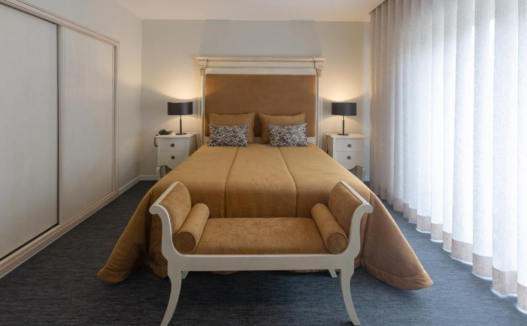 Quarto simples cam cama de casal com colcha marrom clara centralizada, luminárias ao lado da cama e cortina branca e longa. Imagem para ilustrar o post hotéis em Aveiro.