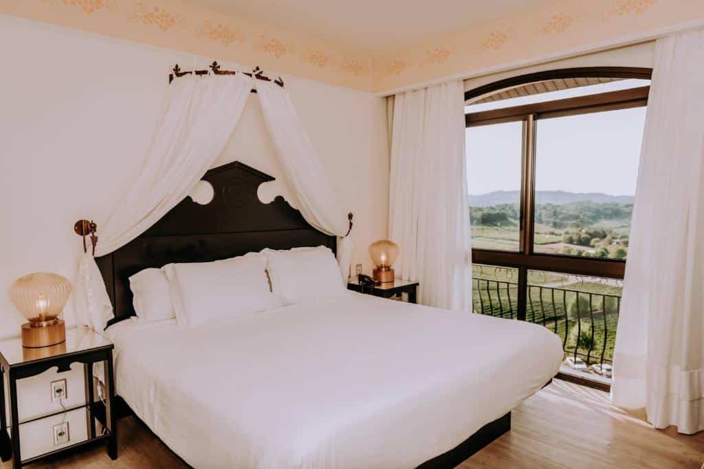 Quarto de casal no Hotel e Spa do Vinho, uma das opções mais icônicas de onde ficar no Vale dos Vinhedos, com cama de lençóis brancos, dossel branco, mesas de cabeceira com abajures e varanda com vista