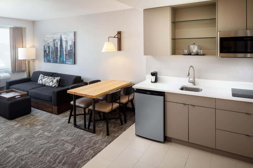 área de estar e cozinha do SpringHill Suites by Marriott com um sofá cama preto, uma mesa de madeira com quatro cadeiras e um balcão de cozinha com uma pia e um frigobar à direita da imagem
