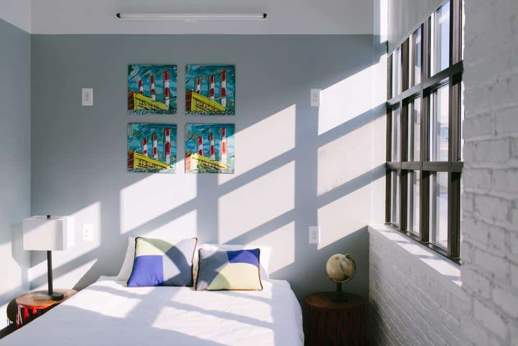 quarto privativo do The Local NY, um dos hostels em Nova York, com uma cama de casal, quatro quadro pequenos em cima da cama, pendurados na parede cinza, e uma janela ampla no lado direito, deixando muita luz solar entrar no ambiente.