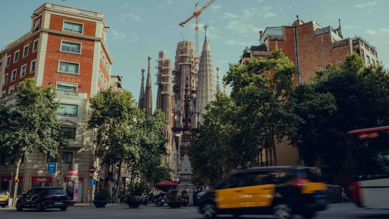 Carros e táxis passando na rua em frente a prédios com a Sagrada Família ao fundo. A foto épara ilustrar o post sobre aluguel de carro em Barcelona, e na rua há muitas árvores. O céu acima está azul. - Foto: Tom Morbey via Unsplash