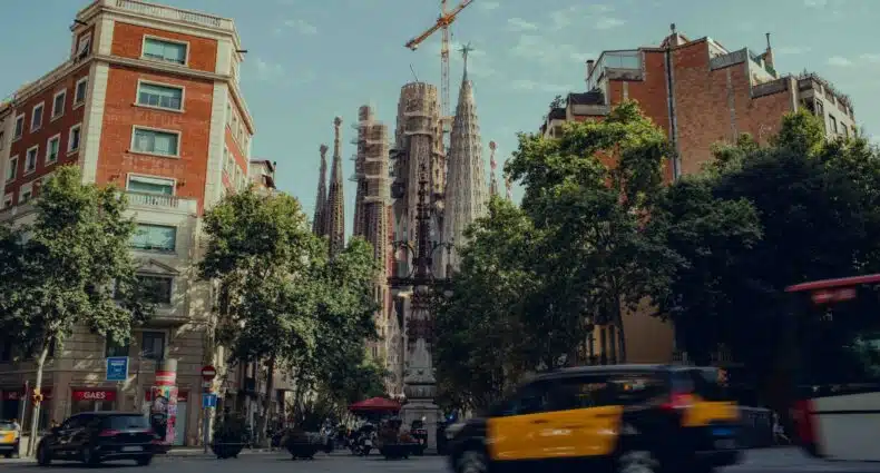 Carros e táxis passando na rua em frente a prédios com a Sagrada Família ao fundo. A foto épara ilustrar o post sobre aluguel de carro em Barcelona, e na rua há muitas árvores. O céu acima está azul. - Foto: Tom Morbey via Unsplash