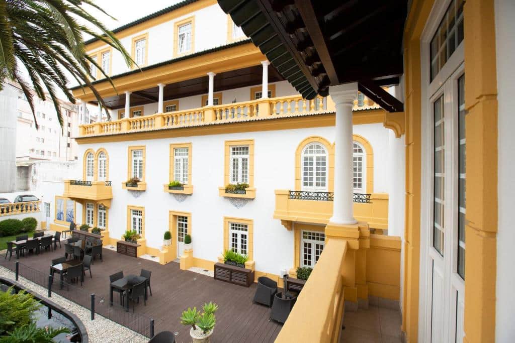 Fachada de hotel com arquitetura clássica, paredes brancas com detalhes em amarelo e deck de madeira com mesas e cadeiras.
