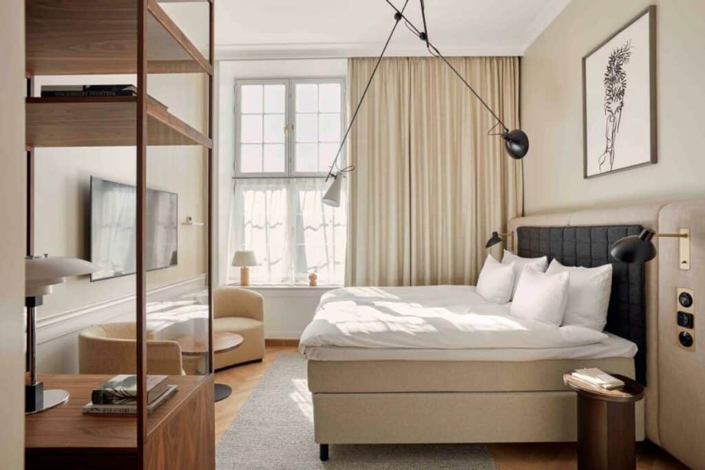 Quarto do hotel Villa Copenhagen com uma cama de casal, armário de prateleiras, mesinha pequena, duas poltronas pequenas, luminárias e uma janela. Foto para ilustrar post sobre hotéis em Copenhague.