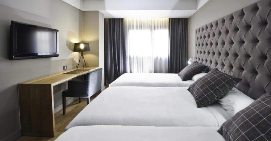 Quarto do Zenit Abeba, uma das recomendações de hotéis baratos em Madri. Três camas encaram uma mesa de trabalho com cadeira, abajur e TV na parede acima. Ao fundo do quarto há uma janela com cortinas cinza e brancas.