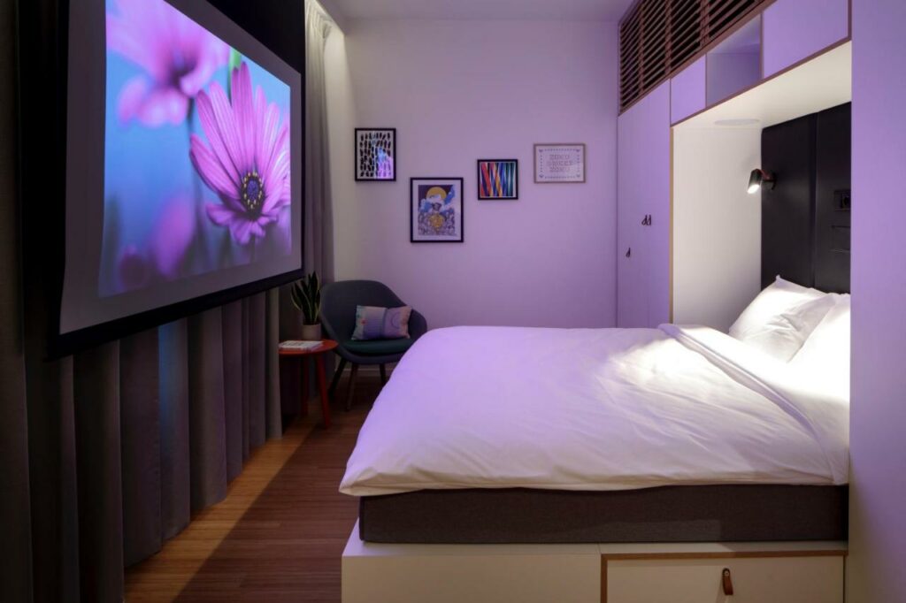 Quarto do hotel Zoku Copenhagen. Há uma cama de casal, ármario integrado na parede, uma cadeira e uma tela de projeção na frente da cortina. O quarto está todo ambientado com iluminação roxa. Foto para ilustrar post hotéis em Copenhague.