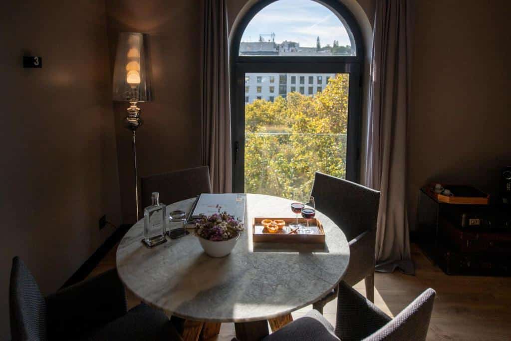 Uma pequena sala com uma mesa redonda com quatro cadeiras estofadas, uma janela com cortinas, com um café da manhã em cima da mesa além de um vaso de flores