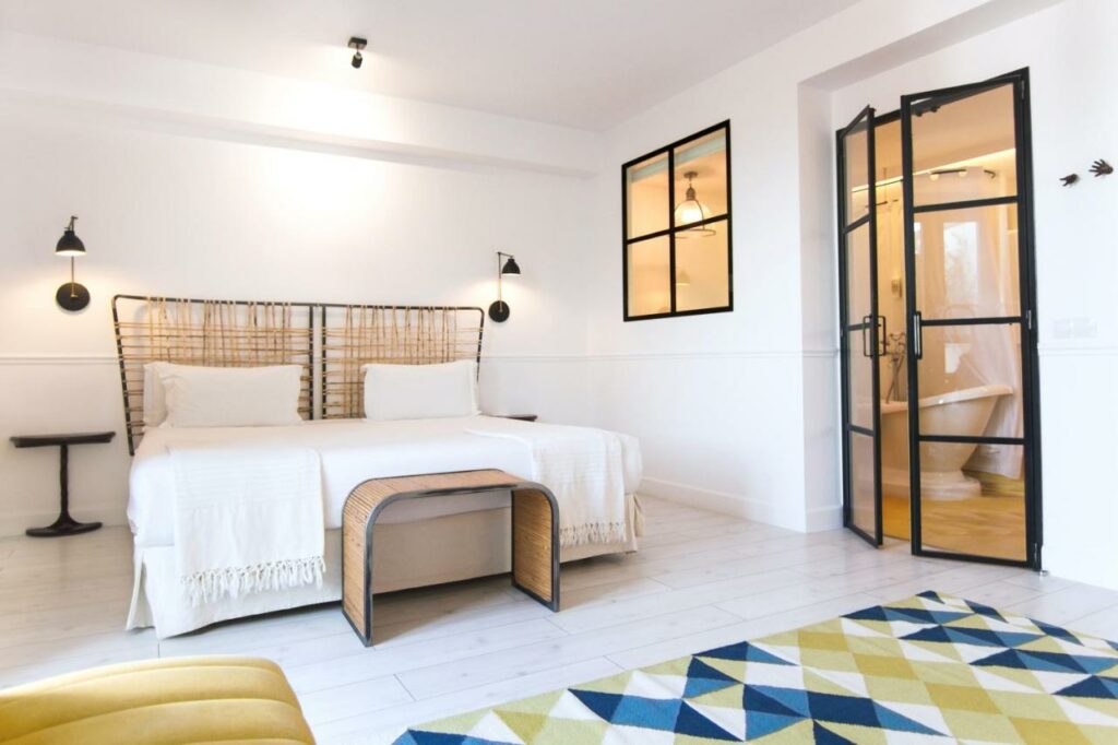 Quarto do 7 Islas Hotel, uma das recomendações de hotéis românticos em Madri. A cama de casal tem jogo de cama branco e mesinhas de cabeceira e luminárias dos dois lados. Um banquinho de madeira está aos pés da cama, e um tapete estampado fica logo a frente. Na parede ao lado há uma janela e uma porta de vidro que leva ao banheiro, onde é possível ver uma banheira.