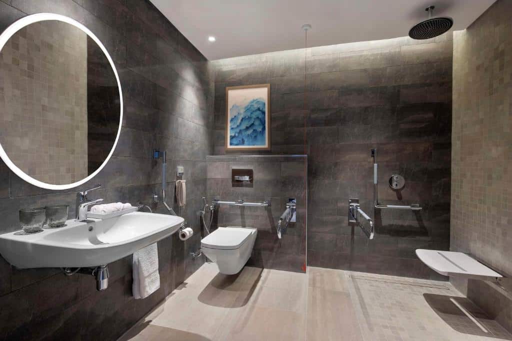 Banheiro acessível do Hilton Abu Dhabi Yas Island. Uma pia do lado esquerdo com dois copos, em cima um espelho redondo. Atrás um vaso sanitário, com um telefone, papel higiênico e barras. Do lado direito o chuveiro, barras e um banco.