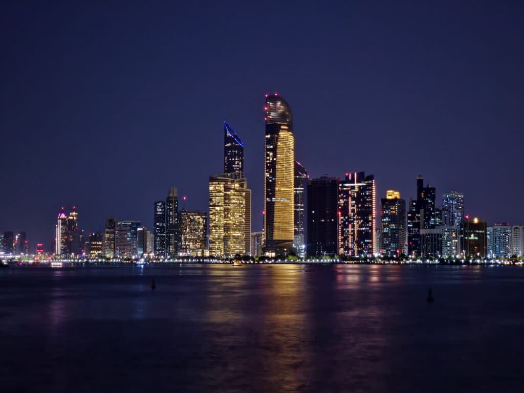 Abu Dhabi a noite. O mar na frente e atrás vários hotéis altos, de diferentes formas, iluminados.
