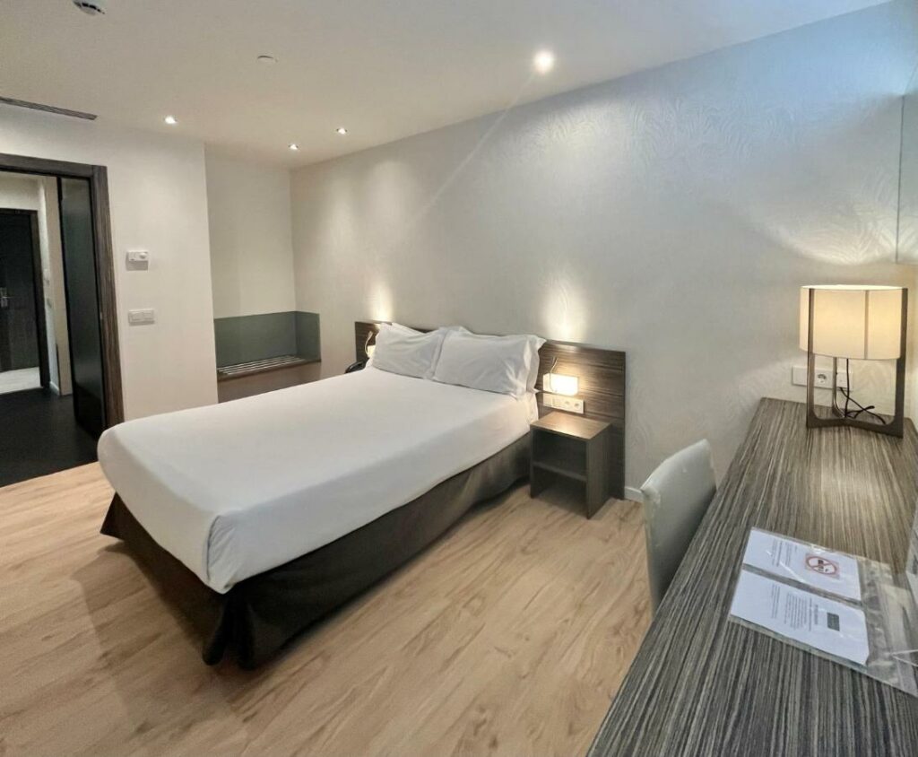 Quarto do Air Rooms Madrid, uma das recomendações de hotéis perto do aeroporto de Madri. A cama de casal tem mesinhas de cabeceira com luminária dos dois lados, e há uma mesa de trabalho com cadeira e abajur na parede ao lado.
