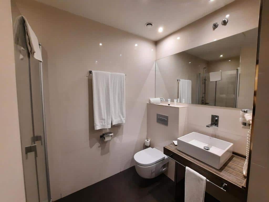 Banheiro do Air Rooms Madrid. A privada branca fica ao lado da pia que tem um secador na parede acima. Um espelho ocupa a parede acima. Em frente está o box com chuveiro e, na parede ao lado, duas toalhas estão penduradas em um suporte acima do papel higiênico.