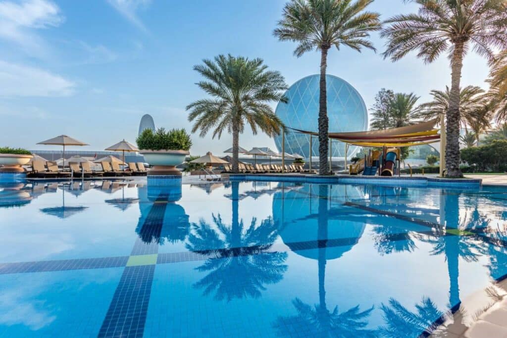 Piscina do Al Raha Beach Hotel. Atrás da piscina, cadeiras de tomar sol, com guarda-sóis e palmeiras. No fundo, no lado direito, um parquinho infantil. No fundo da imagem, uma escultura de vidro, um circulo.