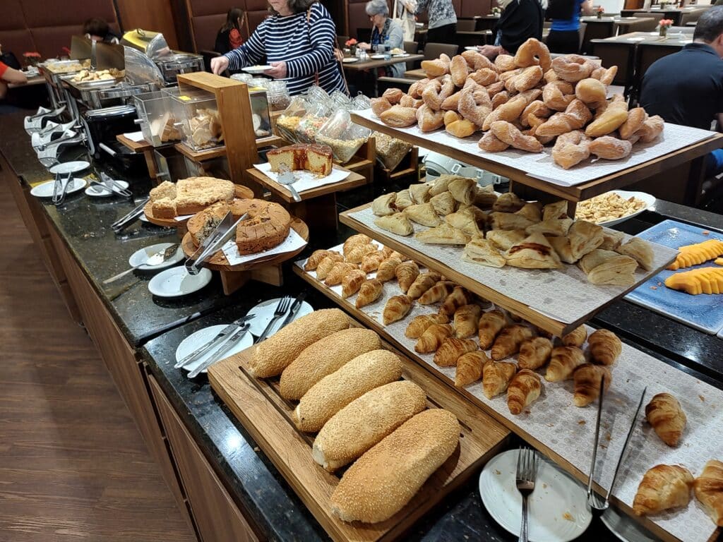 mesa de café da manhã com vários pães, bolos e doces dispostos lado a lado no sistema buffet do hotel. Ao fundo é possível enxergar algumas pessoas sentadas em suas mesas e comendo.