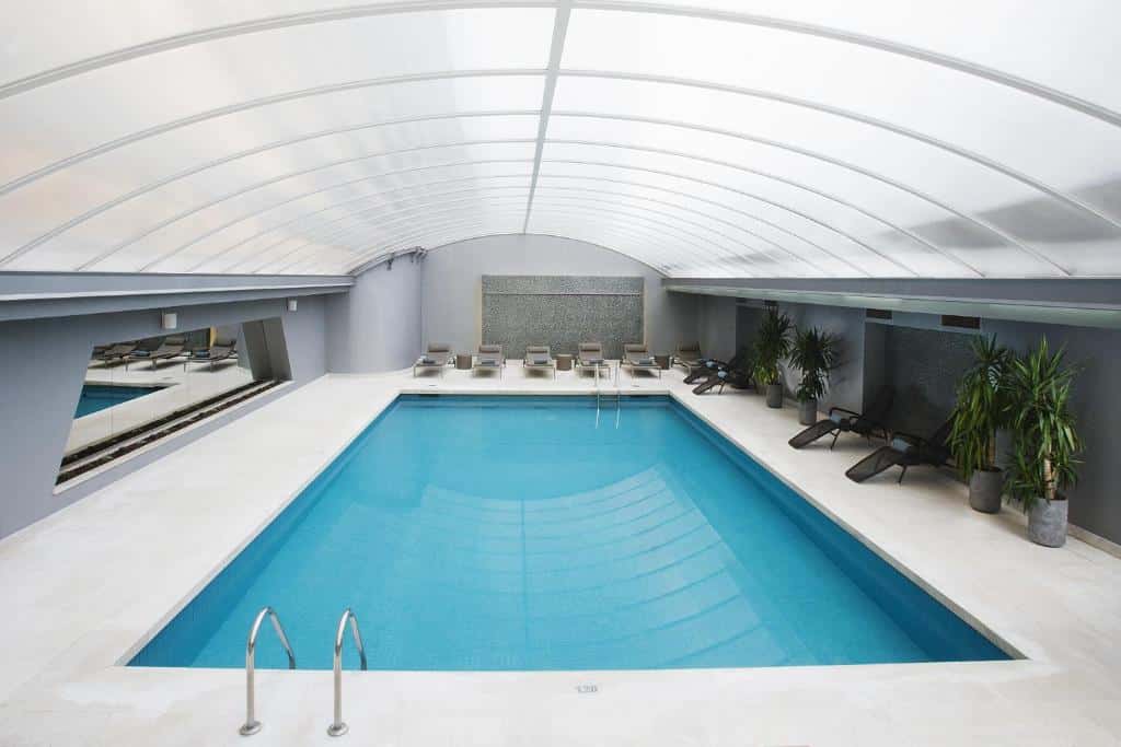 Piscina coberta do Altis Grand Hotel um espaço bem amplo coberto por um telhado transparente, com espreguiçadeiras e vasos de plantas ao redor da piscina