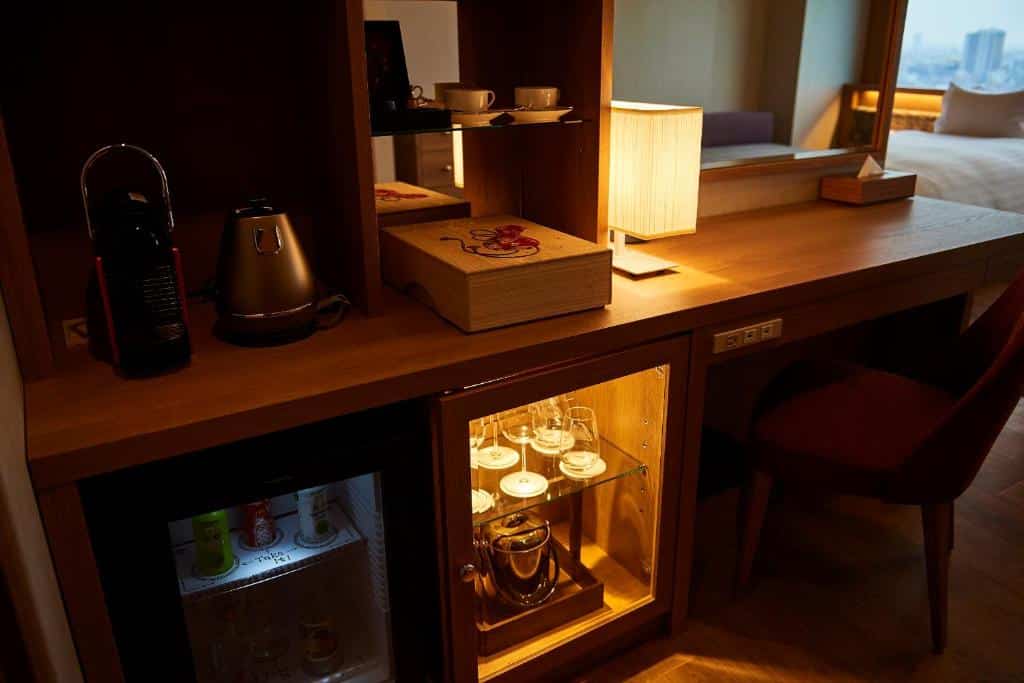Imagem tirada de algumas amenidades em um quarto do Tobu Hotel Levant Tokyo. Há uma mesa de madeira com tomadas, uma cadeira, espelho, abajur, cafeteira, taças, xícaras e bebidas enlatadas.