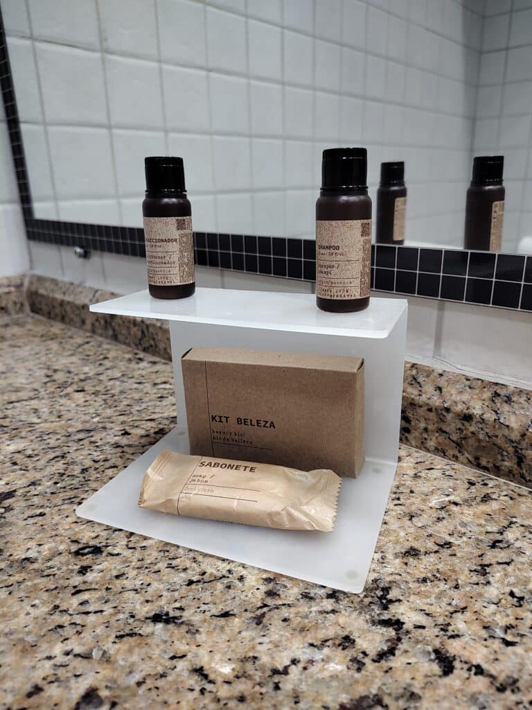 balcão de pedra mostrando o kit de amenidades do banheiro com dois potinhos pequenos de shampoo e condicionados, um sabonete em barra pequeno e um caixinha de papelão com uma toquinha de plástico para banho.