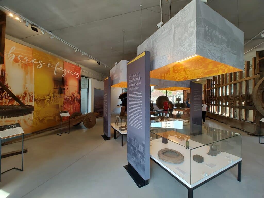 museu interno com vários equipamentos antigos e grandes de madeira dispostos em diferentes locais. Há também pequenas mesas de exposição, cobertas com caixas de vidro, onde há vários objetos pequenos.