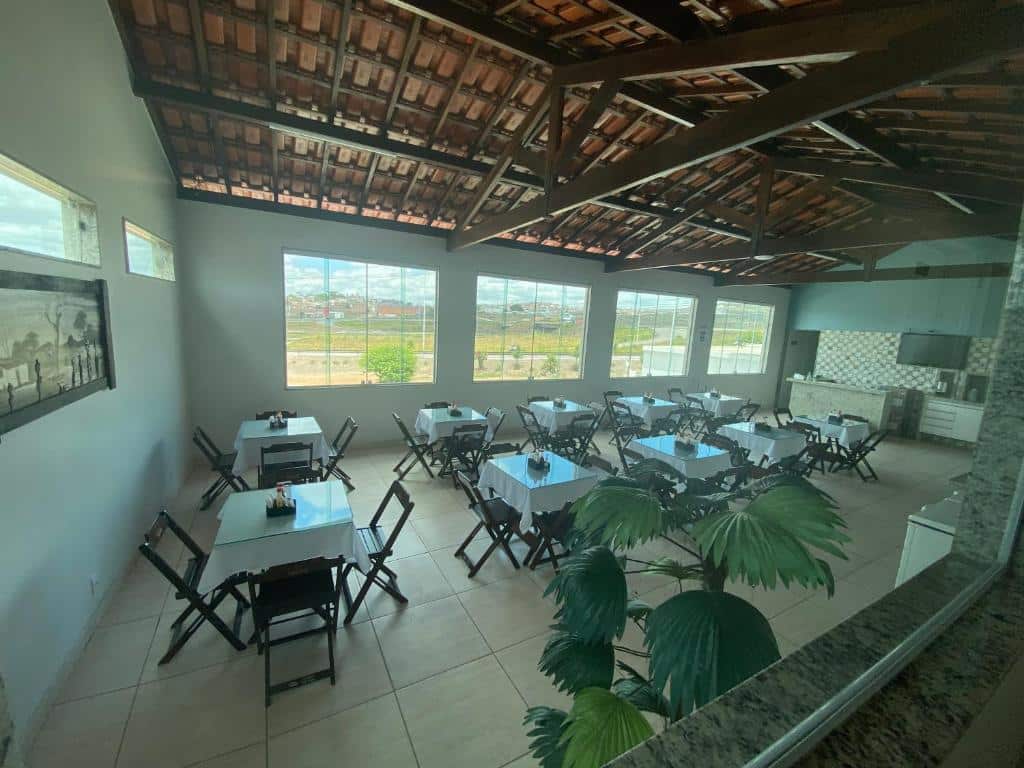 Área para refeições da Pousada Trevo Caruaru. O saguão é amplo, possui diversas mesas e cadeiras, ao fundo há quatro janelas grandes e no lado direito há um balcão com uma TV presa a parede.