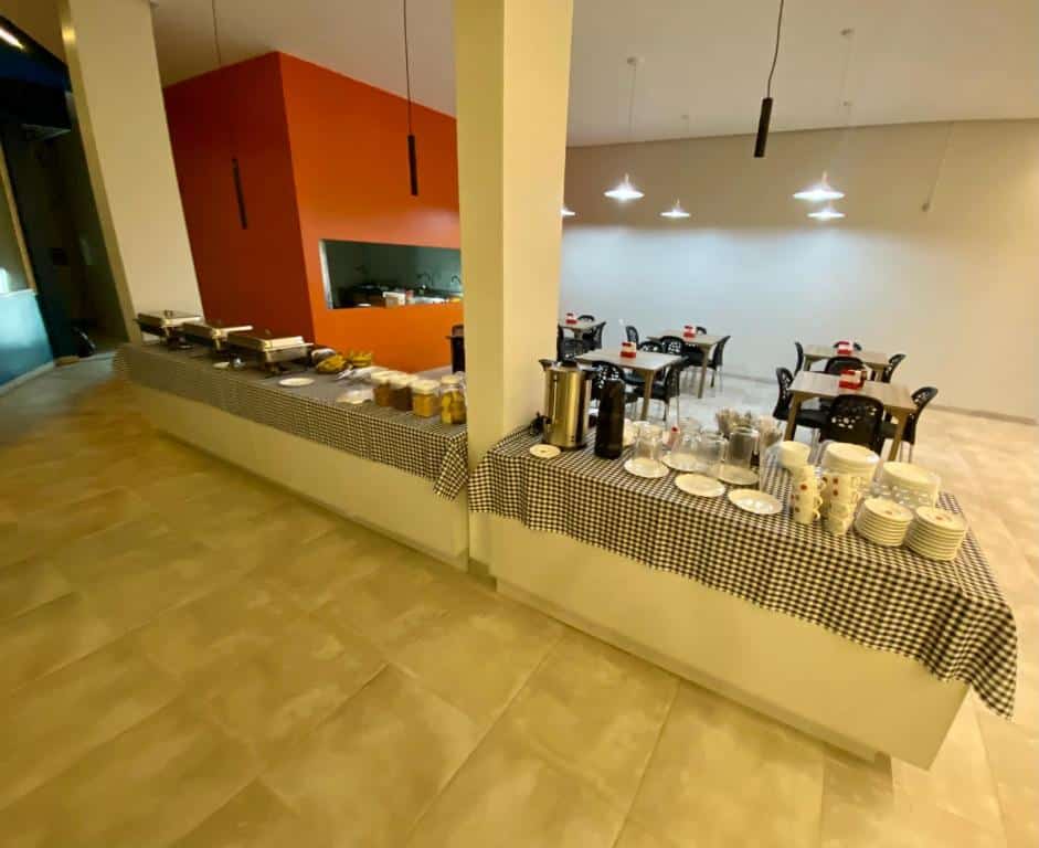 Área para refeições do Rota 22 hotel Caruaru. Em cima de uma longa bancada estão pratos, xícaras, talheres, garrafas térmicas, potes e panelas. Ao fundo estão diversas mesas com cadeiras e ao lado esquerdo é possível ver um pouco da cozinha.