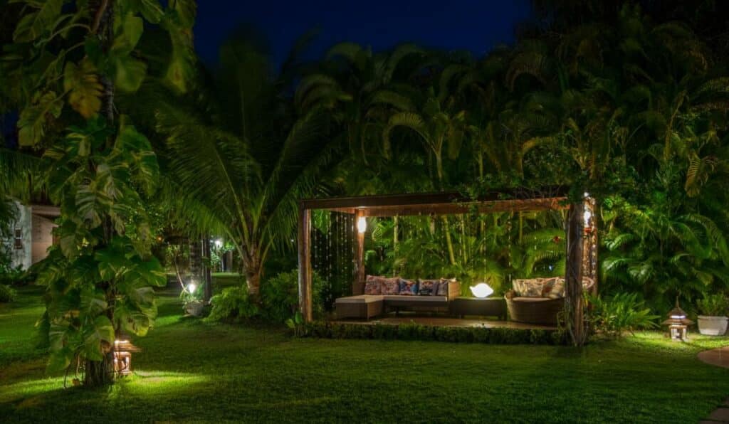 Jardim da Pousada Villa Cumuru. Árvores iluminadas ao redor de um de deck iluminado com cadeiras e almofadas.
