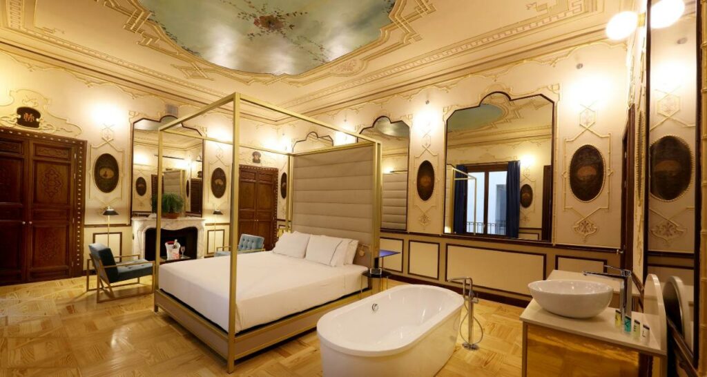 Quarto do Axel Hotel Madrid - Adults Only, uma das recomendações de hotéis românticos em Madri. No centro do lugar está uma cama de casal com dossel e mesinhas dos dois lados. Ao lado direito há uma banheira em frente à pia com espelho e amenidades de banho. Já do lado esquerdo fica uma área de estar com poltronas e mesa de centro. As paredes e o teto tem um estilo renascentista, e há grandes espelhos em todas as paredes.