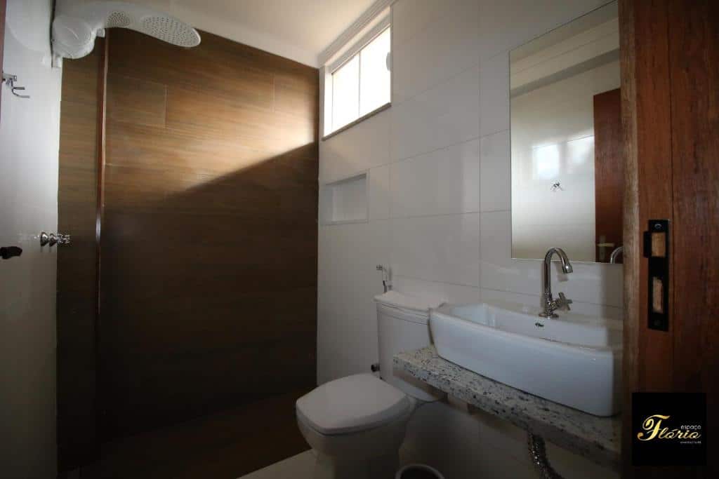 Banheiro do Espaço Flório Hotel. Há uma bancada de mármore pequena com uma pia em cima e um espelho, do lado está o vaso sanitário e um espaço para o chuveiro.